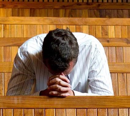 Fotografía de un varón vestido de camisa blanca sentado en el banco de una iglesia inclinada su cabeza sobre sus manos con los dedos entrelazados en el acto de orar.