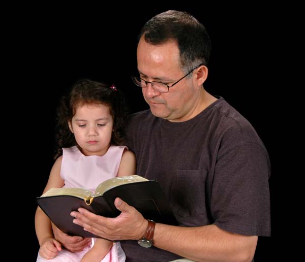 En esta fotografía un padre sostiene una Biblia abierta en su mano frente a su hijita sentada en su falda, ilustración para el tema Las iglesias de Cristo os saludan, en iglesia-de-cristo.